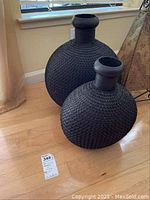 Two black vintage chainmail vases placed on a wooden floor by a window, showing the spherical mesh design and narrow neck openings, with visible wear.