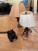 Three artistic lamps shown together: one with sculptural metal ball base and trapezoidal lampshade, one with ceramic bear base and tan stitched lampshade, and one with dark animal base and cream lampshade.