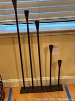 Five black steel candle holders of varying heights standing on the floor against a wall, shown as a group illustrating size and style.