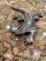 Steel cast lizard sculpture showing detailed legs, toes, eyes and tail on a granite surface.