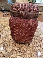 Front view of one of three woven baskets from Bali, showing rounded square shape, woven texture, and lid with strip detail.