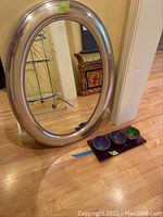 Full view of oval silver wood frame mirror leaning against wall and set of three painted artistic bowls on purple rectangular tray on the floor.