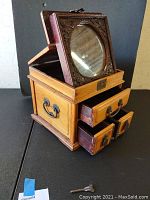 View of the early 1900s Asian bamboo jewelry box partially open showing carved wood frame around oval mirror, top locking mechanism, three front drawers with metal handles, and a small metal key on the surface in front of it.