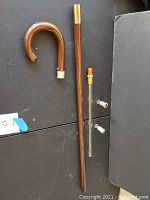 Photo showing the disassembled wooden cane with curved handle, cane shaft, glass decanter tube and cork stopper, and two small glass shot glasses laid out on black surface.