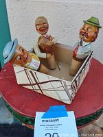 Three wood-carved mechanical bottle stoppers in a cardboard box displayed on a round stool