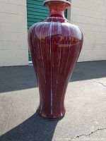 Front view of the tall Asian porcelain vase with sang de boeuf glaze and white drip streaks, shown outdoors on pavement against a brick wall.