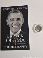 Front cover of the paperback biography book titled 'Barack Obama The Biography' and the 44th President commemorative coin in a protective capsule