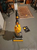 Front view of yellow and gray upright Dyson vacuum on concrete floor with power cord trailing.