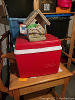 Red Rubbermaid cooler with white lid seen on wooden table with hand-painted wooden birdhouse on top.