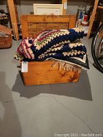 Wood chest with multicolor crochet blanket draped over, chest shows scratched surface and honey-brown finish