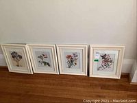 Four cream-colored framed botanical prints leaning against a wall on wooden floor