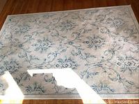 Full view of the worn acrylic rug with floral and vine designs in blue and gray tones on a beige background, laid on wooden floor.
