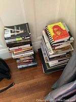 Two stacks of assorted books leaning against wall on wood floor, showing several titles and different book sizes.