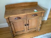 Front angled view of antique pine cabinet showing two drawers with carved wooden handles, two doors with black knobs, ornate side columns, and raised backboard.