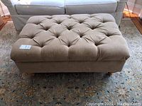 Side view of a beige tufted ottoman placed in front of a light colored couch on a patterned area rug.