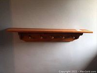 Wooden wall-mounted shelf with five round wooden hooks visible beneath, mounted on a plain wall.