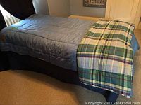 Side view of single bed with light blue comforter and plaid blanket folded at foot of bed, showing bed on carpeted floor with window and dark curtains beside.