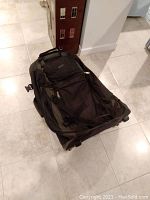 Photo of top and side view of black wheeled backpack with multiple compartments, placed on tiled floor near wall with dollhouse present.