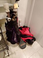 Photo of black and purple golf bags filled with multiple golf clubs and a red and black jacket on floor