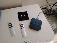 Photo of Apple TV device, two remotes and setup guide on white table