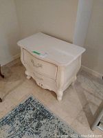 Full view of white wooden night stand with distressed finish and two dovetailed drawers showing ornate handles and curved details.