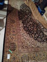 Full view of rectangular rug laid out on concrete floor, partial roll at one end, surrounding storage items visible.