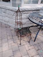 Photo showing metal trellis and glass-top patio end table on outdoor patio pavers with a stone wall background.