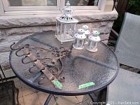 Photo showing large ornate metal wall hook and four white metal candle lanterns on glass-top outdoor table