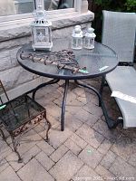Metal and glass patio table shown outdoors on a stone patio, alongside a lounge chair and small decorative lanterns on top, showcasing the table's condition and materials.