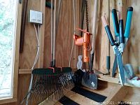 Wide view showing metal garden rake with black tines and green handle, a garden edger with green handle, and two shovels with wooden handles against wood wall.