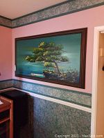 Framed oil painting of a tree landscape with water body and rocky terrain.