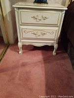 Front view of one cream-colored, footed nightstand with two drawers, gold-tone handles, and decorative gold accents on drawers and edges.