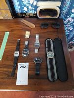 View of six watches: two analog and four digital on a wooden surface along with sunglasses, shoehorn, and money clip.
