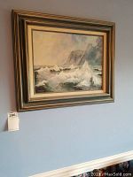 Framed original acrylic painting of a seascape showing waves breaking against cliffs, hung on light blue wall with lot tag.