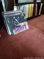 Stack of vinyl records with Isaac Hayes Shaft album in front, set on red carpet by shelving
