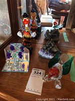 Colorful hand-blown glass clown figurine, glass bird with green wings, amethyst cluster with mining scene, and mirrored grape motif piece arranged on wooden surface in front of mirror.