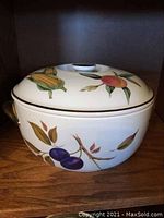 Front view of porcelain casserole dish with painted fruit and vegetable designs and gold-tone handles on lid and sides.