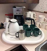 Full view of coffee maker with glass carafe, white electric kettle, white carafe with lid, six green mugs, three white mugs stacked, metal tea strainer, and oval tray on countertop.