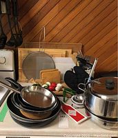 Overview of frying pans, egg poacher, cutting boards, splatter screen, wooden spoon rests, and oven mitt on countertop with wood paneling background