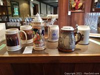 Front view of the 6 beer steins and mugs arranged on wood surface showing variety in types, styles, and sizes.