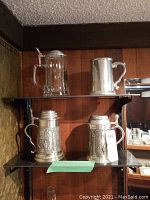 Four beer steins displayed on a shelving unit, including one glass with pewter lid and three pewter steins with embossed scenes.