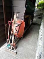 Photo showing orange metal wheelbarrow with black handles and several garden tools including rakes, brooms, and hoes leaning against the wall.