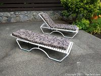 View of two white metal frame chaise lounges with floral cushions placed outdoors on a concrete surface, showing full chairs in reclined and upright positions.
