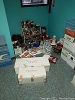 Photo showing assorted Christmas decorations including ornaments displayed on a tiered rack, multiple boxes and plastic containers, and Santa and Mrs. Claus doll boxes on the floor.