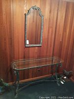 Full view of the metal framed glass top table with matching decorative mirror hanging above it against wood-paneled wall.