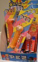Photo showing seven PEZ candy dispensers in original blister packaging inside a blue PEZ brand display box. Various colorful dispenser heads including a whistle version are visible.
