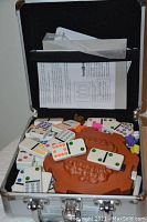 Open metal storage case showing plastic domino tiles with colored dots, brown plastic train-shaped game base, colorful small game pieces, and instruction sheets inside.
