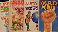 Four Mad Magazine books standing upright showing covers with titles including 'Mad's Brain Ticklers,' 'Don Martin Cooks Up More Tales,' 'Mad's Looks At Our Sick World,' and 'Mad Power' with a fist on the cover.