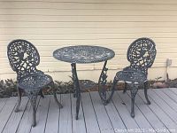 Full view of black metal outdoor table and two matching chairs with detailed cut-out designs and some paint chipping.