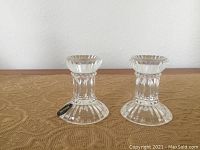 Pair of clear crystal candle sticks with fluted design on base and stem. Both have rounded, wide bases and shorter stems feeding into the candle cup holders. Clean and shiny condition.